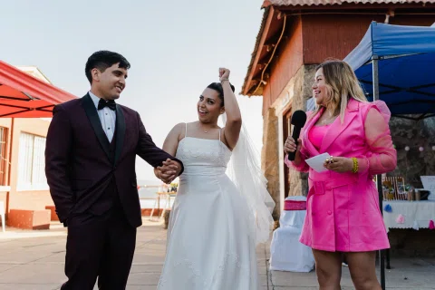 Hombre y mujer con vestido blanco están tomados de la mano junto a una mujer vestida de rosado que sostiene un micrófono.