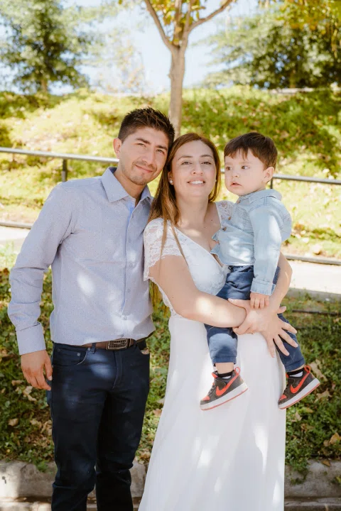 Hombre con camisa celeste y mujer con vestido blanco posan junto a un niño al aire libre.