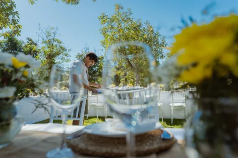 Hombre prepara una mesa exterior visto entre copas y flores desenfocadas.