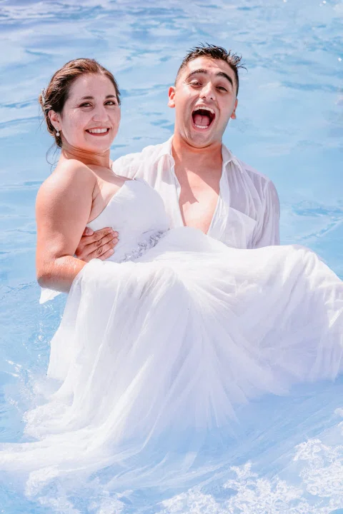 Hombre sosteniendo a la mujer con vestido blanco dentro de una piscina.