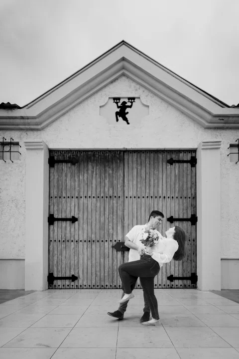Hombre sostiene inclinada hacia atrás a una mujer con ramo frente a un portón de madera, en blanco y negro.