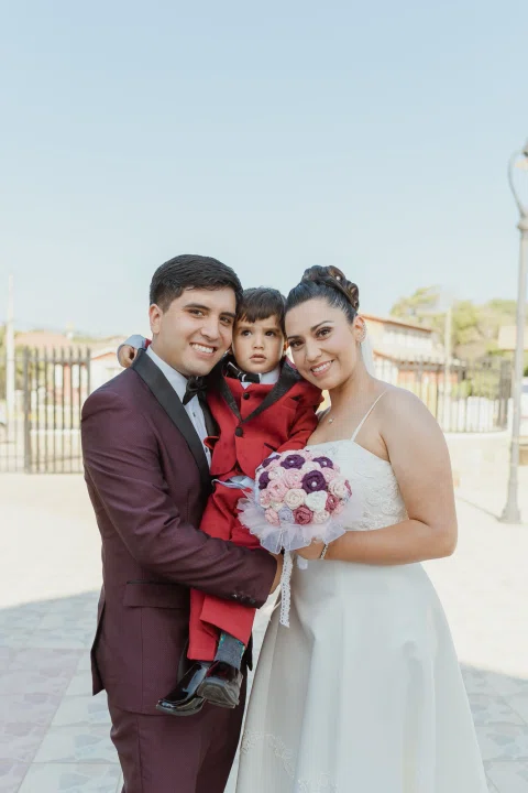 Hombre sostiene en brazos a un niño vestido de rojo junto a una mujer con vestido blanco que lleva un ramo rosado.