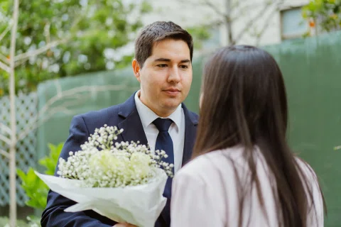 Hombre con traje oscuro sostiene un ramo de flores blancas frente a una mujer de chaqueta clara.