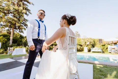 Hombre con camisa blanca y tirantes azules permanece de pie frente a una mujer con vestido blanco sentada en una silla sobre la pista.