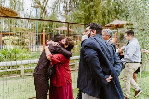 Hombre con traje oscuro abraza a una mujer con vestido rojo mientras varias personas permanecen alrededor.