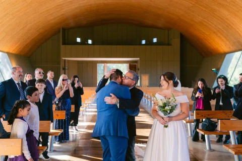 Hombre con traje azul abraza a un hombre mayor mientras una mujer con vestido blanco y ramo permanece al lado.