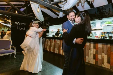 Hombre con traje azul abraza a una mujer vestida de negro frente a una barra; al fondo, una mujer con vestido blanco sostiene a una niña.