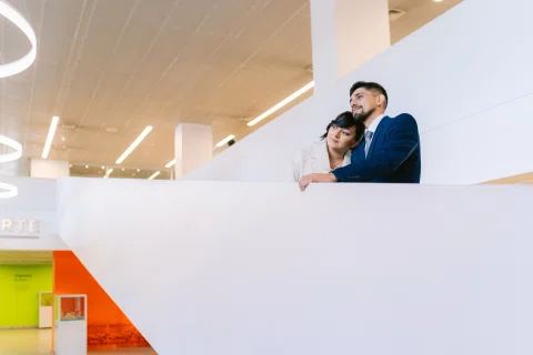 Hombre con traje azul abraza a una mujer con traje claro junto a un muro blanco geométrico.