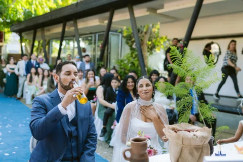 Hombre con traje azul bebe de un vaso junto a una mujer con vestido blanco sentada a su lado.