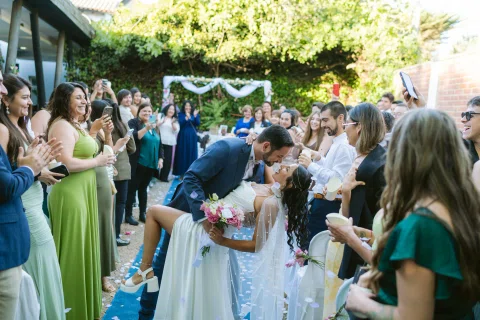Hombre con traje azul besa a una mujer con vestido blanco mientras la inclina hacia atrás entre personas que aplauden.