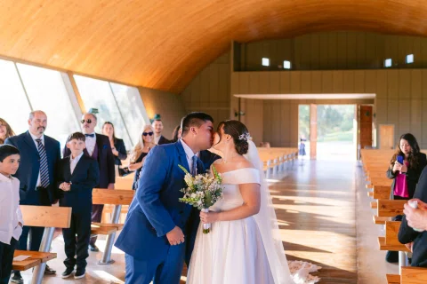 Hombre con traje azul besa a una mujer con vestido blanco y ramo dentro de una capilla.