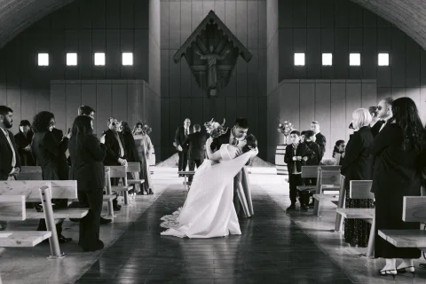 Hombre con traje azul besa y sostiene inclinada a una mujer con vestido blanco en el pasillo, en blanco y negro.
