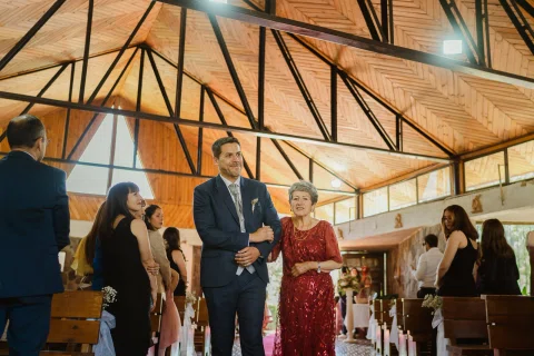 Hombre con traje azul camina del brazo de una mujer con vestido rojo por el pasillo de la iglesia.