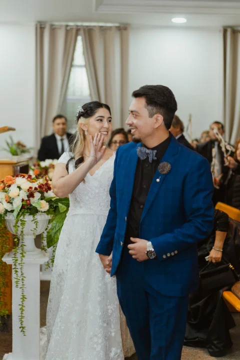 Hombre con traje azul camina junto a una mujer con vestido blanco que levanta una mano en un saludo.