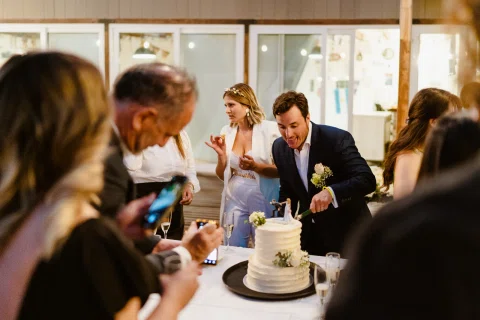 Hombre con traje azul corta una torta blanca mientras una mujer vestida de blanco está a su lado comiendo un bocado.