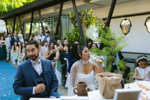 Hombre con traje azul permanece de pie junto a una mujer con vestido blanco sentada; una niña aparece al fondo.
