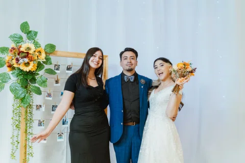 Hombre con traje azul posa entre dos mujeres frente a un fondo claro con decoración floral.