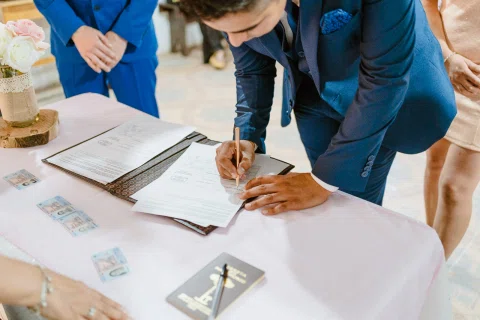 Hombre con traje azul inclinándose para firmar un documento sobre una mesa.