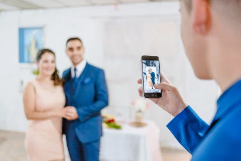 Hombre con traje azul fotografía con su teléfono a un hombre y una mujer que posan al fondo.