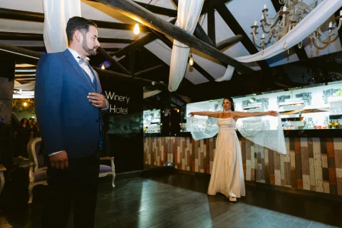 Hombre con traje azul permanece frente a una barra mientras una mujer con vestido claro extiende los brazos detrás.