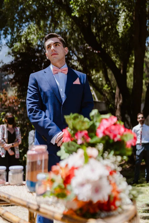 Hombre con traje azul permanece junto a una mesa con flores en primer plano.