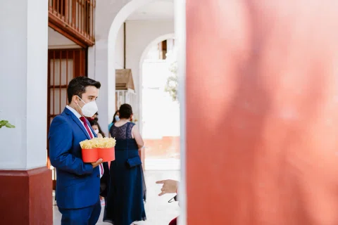 Hombre con traje azul y mascarilla sostiene un recipiente rojo junto a la entrada de la iglesia.
