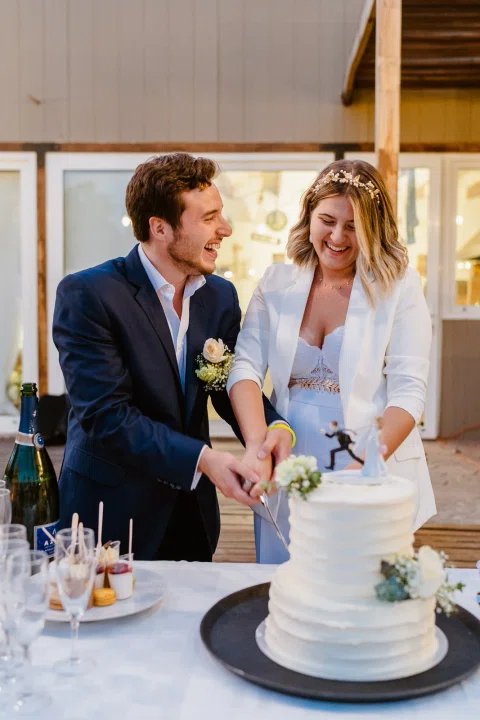 Hombre con traje azul y mujer vestida de blanco cortan juntos una torta blanca con un cuchillo.