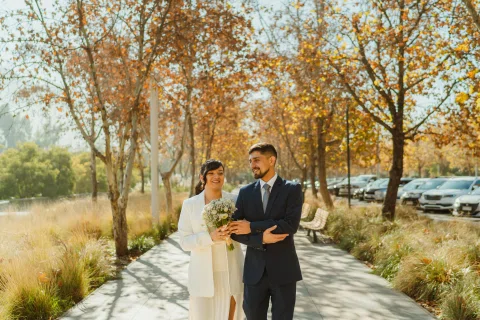 Hombre con traje azul y mujer con traje claro y ramo caminan juntos por un parque de tonos otoñales.