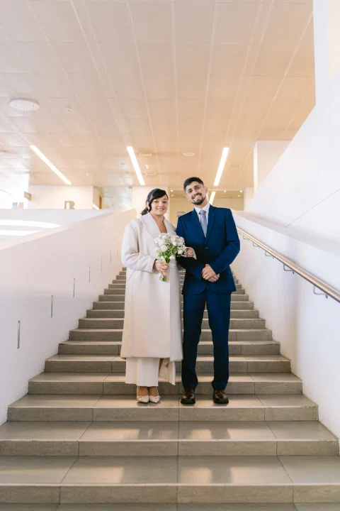 Hombre con traje azul y mujer con traje blanco posan juntos en una escalera interior.