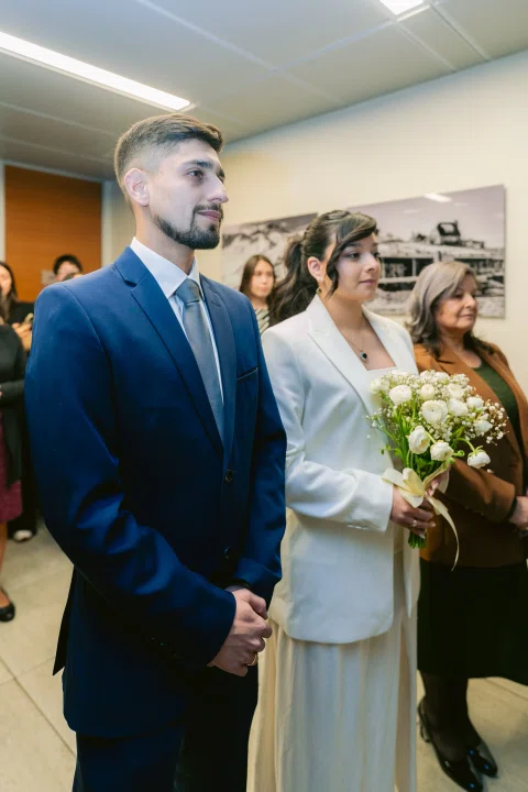 Hombre con traje azul permanece junto a una mujer con traje blanco que sostiene un ramo.