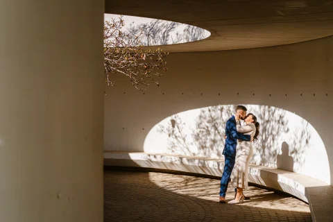 Hombre con traje azul y mujer con traje claro se abrazan bajo una abertura curva iluminada por luz lateral.
