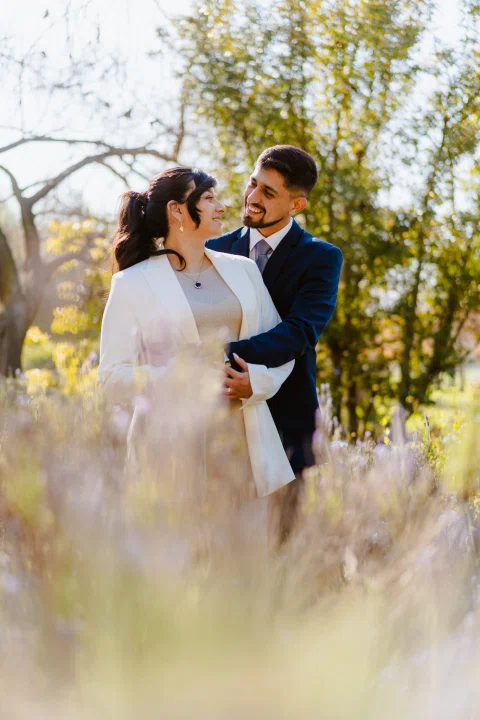 Hombre con traje azul y mujer con traje claro se abrazan detrás de flores altas desenfocadas.