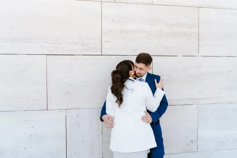 Hombre con traje azul y mujer con traje claro se abrazan frente a un muro de piedra clara.