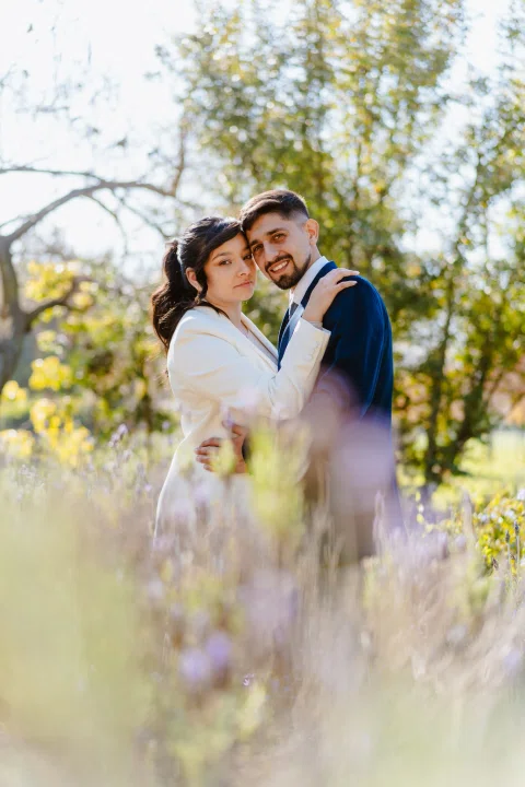 Hombre con traje azul y mujer con traje claro permanecen abrazados entre flores y vegetación desenfocada.