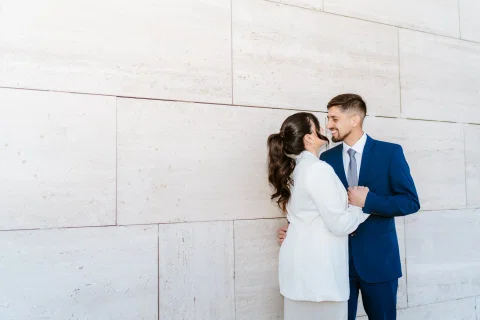 Hombre con traje azul y mujer con traje claro permanecen juntos y sonríen frente a un muro de piedra clara.