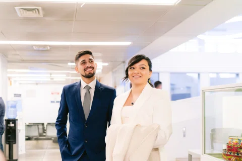 Hombre con traje azul y mujer con traje claro posan juntos dentro de una sala.