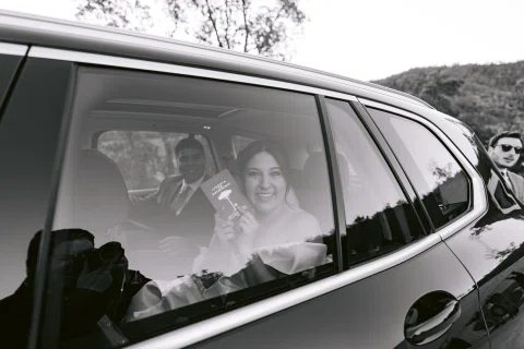 Hombre con traje azul y mujer con vestido blanco aparecen dentro de un automóvil vistos a través de la ventana, en blanco y negro.