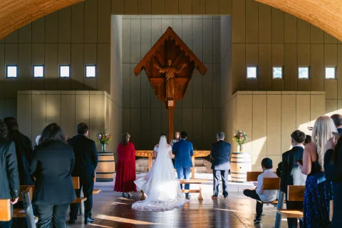 Hombre con traje azul y mujer con vestido blanco avanzan hacia el frente de una capilla vistos desde atrás.