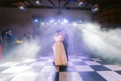 Hombre con traje azul y mujer con vestido blanco bailan abrazados bajo luces y humo.