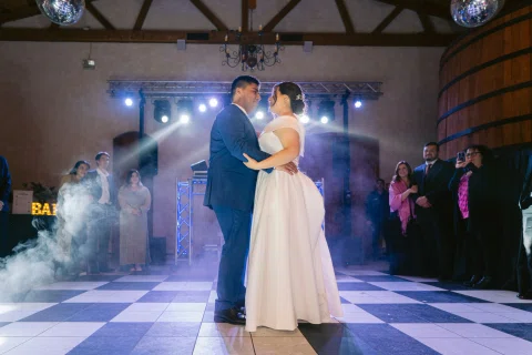 Hombre con traje azul y mujer con vestido blanco bailan juntos en el centro de una pista con humo escénico.