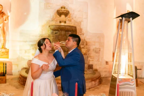 Hombre con traje azul y mujer con vestido blanco beben de copas altas con los brazos entrelazados.