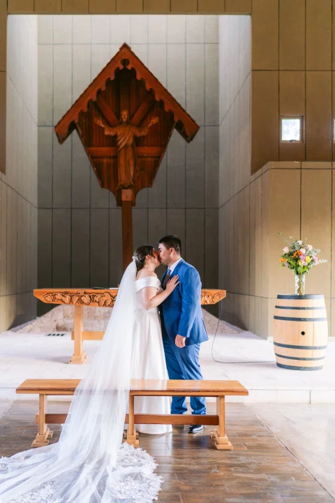 Hombre con traje azul y mujer con vestido blanco se besan frente al altar.