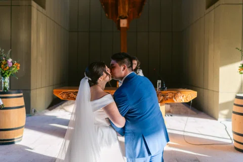 Hombre con traje azul y mujer con vestido blanco se besan frente a una mesa de madera.