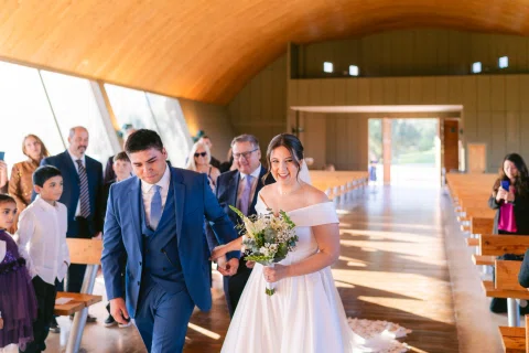 Hombre con traje azul y mujer con vestido blanco caminan tomados de la mano por el pasillo.