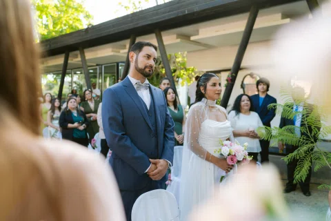 Hombre con traje azul y mujer con vestido blanco permanecen de pie uno junto al otro al aire libre.
