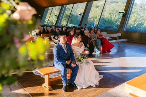 Hombre con traje azul y mujer con vestido blanco están sentados juntos, vistos entre flores y personas al fondo.
