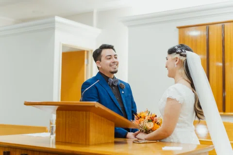 Hombre con traje azul y mujer con vestido blanco se miran de pie frente a un atril de madera.