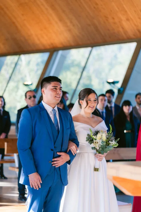 Hombre con traje azul y mujer con vestido blanco permanecen juntos dentro de la capilla.