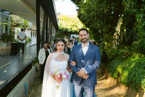 Hombre con traje azul y mujer con vestido blanco posan juntos al aire libre.