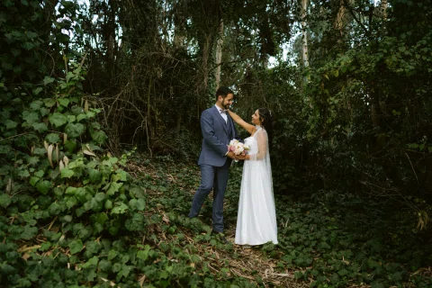 Hombre con traje azul y mujer con vestido blanco posan juntos entre vegetación.